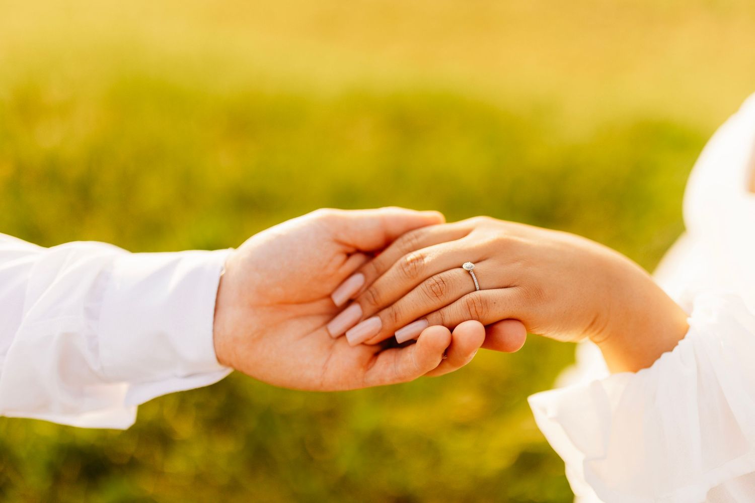 Close-up of couple holding hands outdoors, woman wearing diamond engagement ring, golden bokeh background.