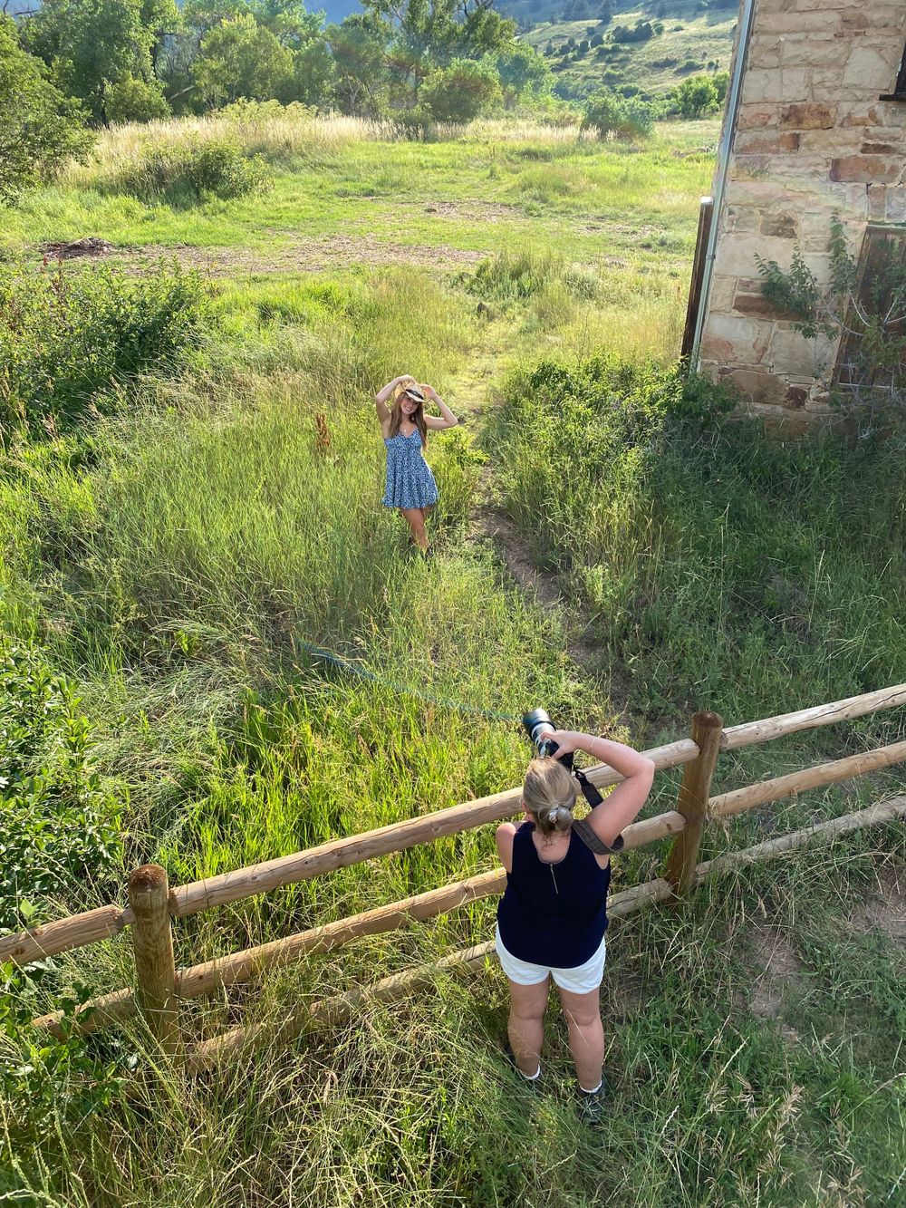 People taking photos in grassy field near rustic wooden fence with mountains in background.