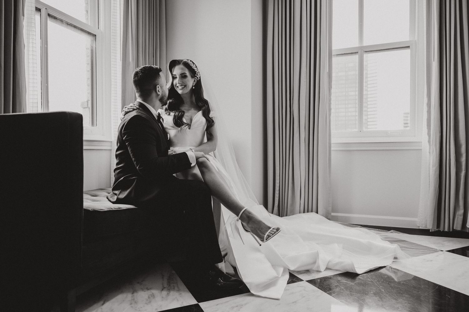 A black and white wedding photo by a window with natural light streaming in.