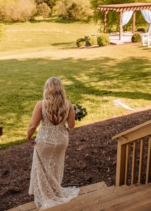 A lace wedding dress flows elegantly as someone walks down wooden stairs toward a garden gazebo.