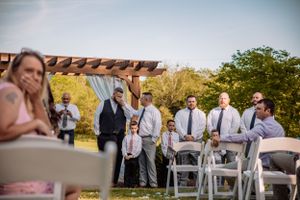 Wedding guests seated in white chairs under trees during an outdoor ceremony.