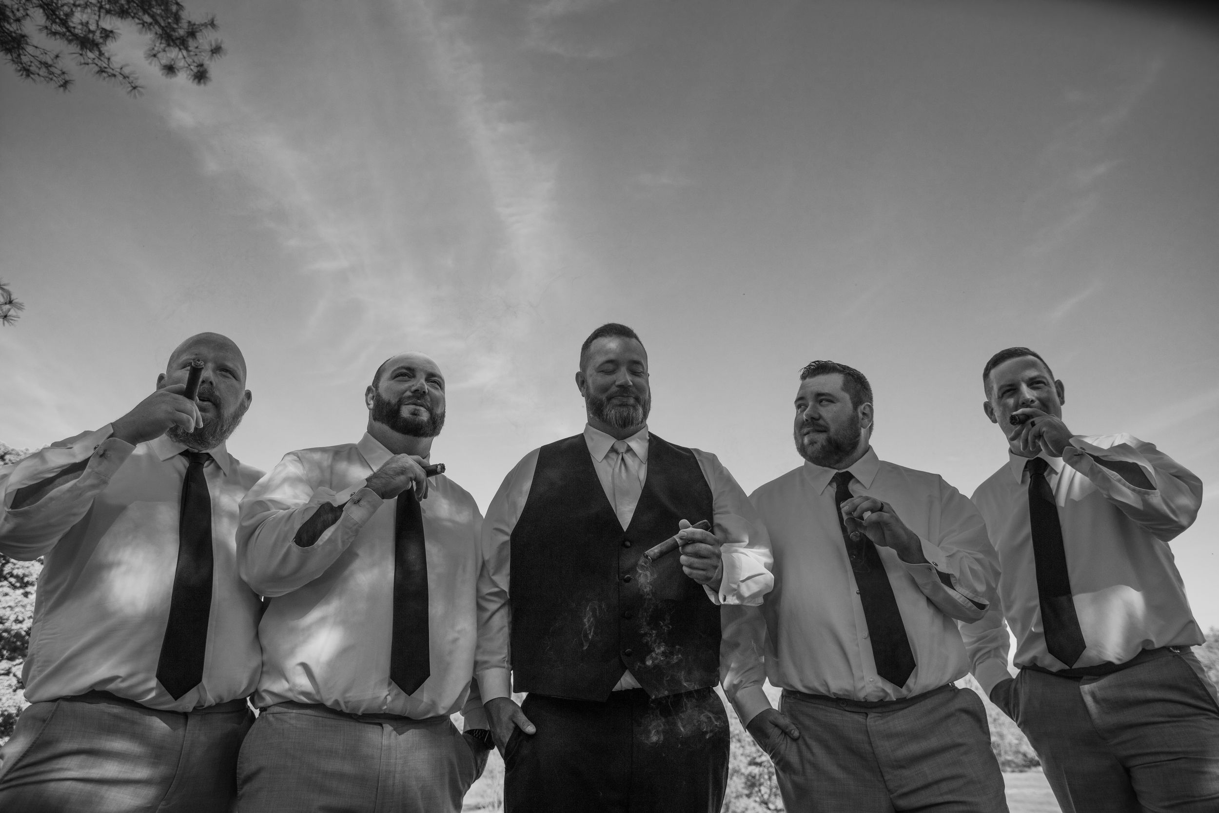Black and white photo of groomsmen posing together outdoors against a bright sky.