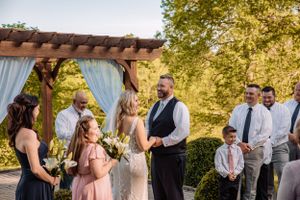Outdoor wedding ceremony under wooden pergola with blue drapes and natural landscaping.