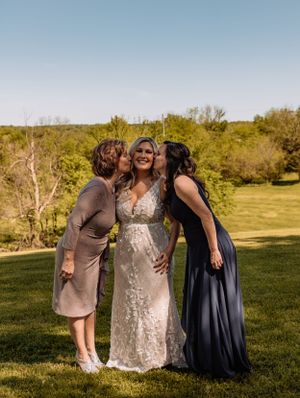 Three people share a tender moment on a grassy hillside during a wedding celebration.