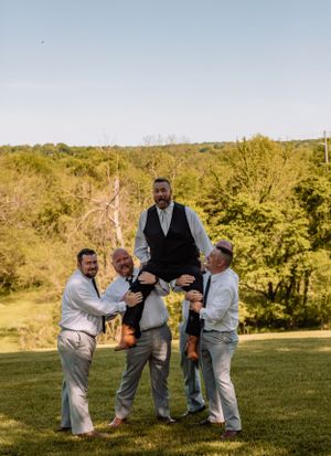 Groomsmen playfully lift someone during outdoor wedding photos on a grassy hill.