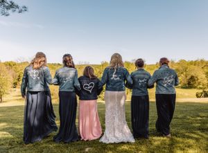 Wedding party wearing matching denim jackets stands with backs to camera on grassy hill.