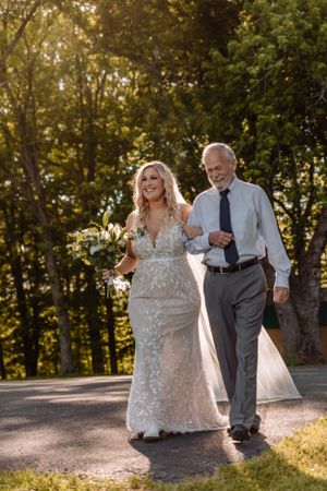 Wedding procession walking down a sunlit outdoor path surrounded by autumn trees.