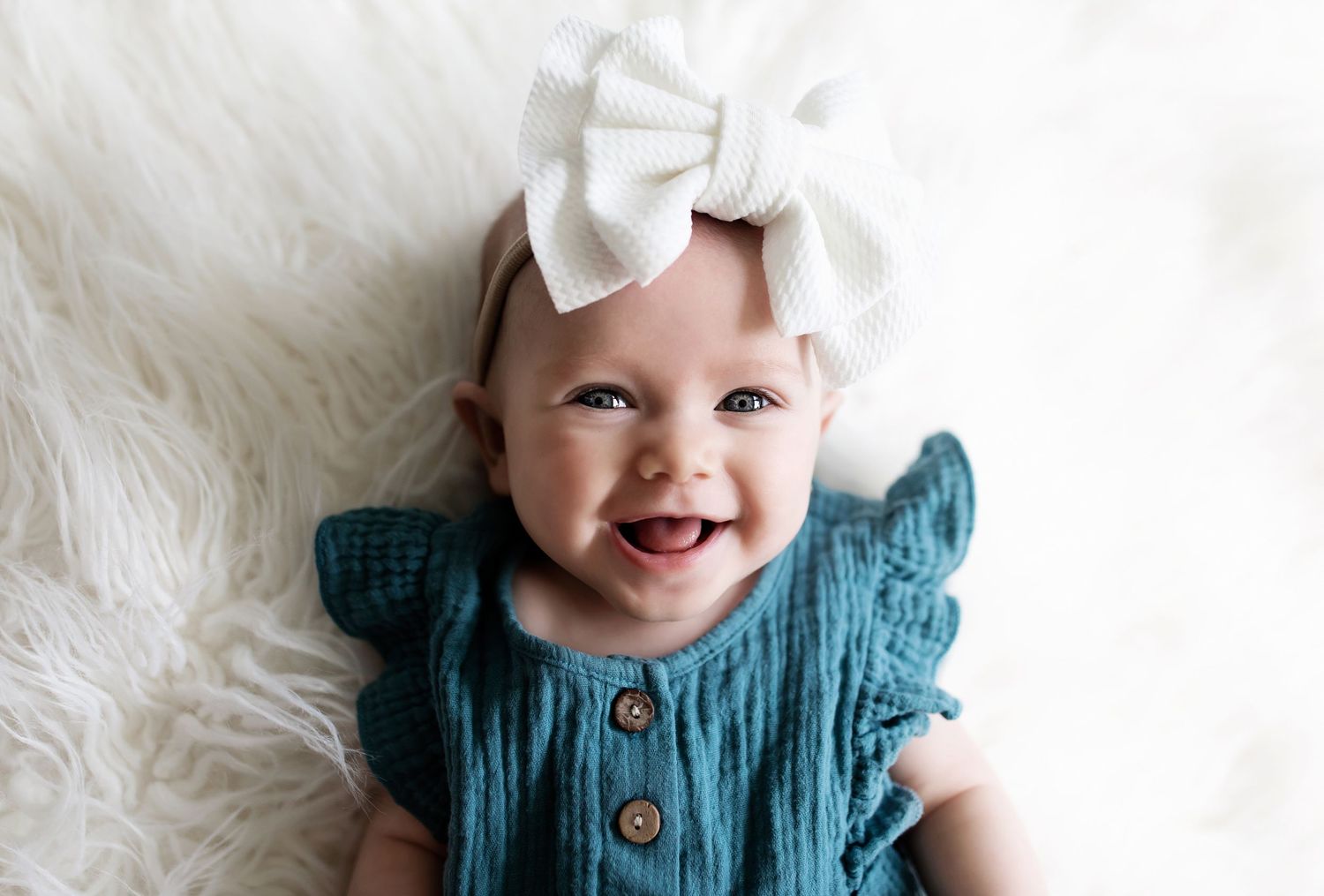A baby wearing a teal dress outfit lies on fluffy white fabric.