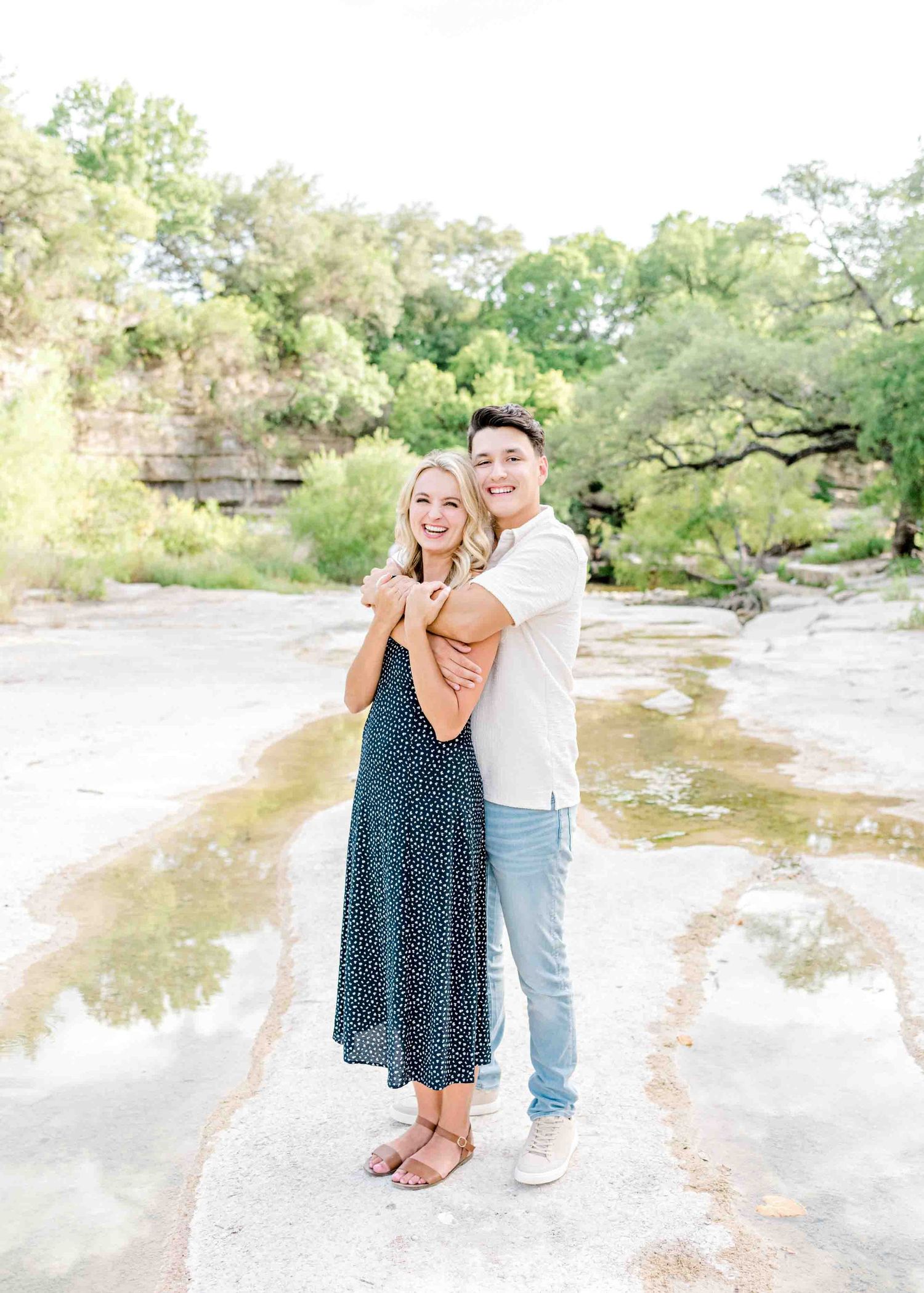 Romantic couple embracing by a scenic waterfront with natural rock formations and greenery.