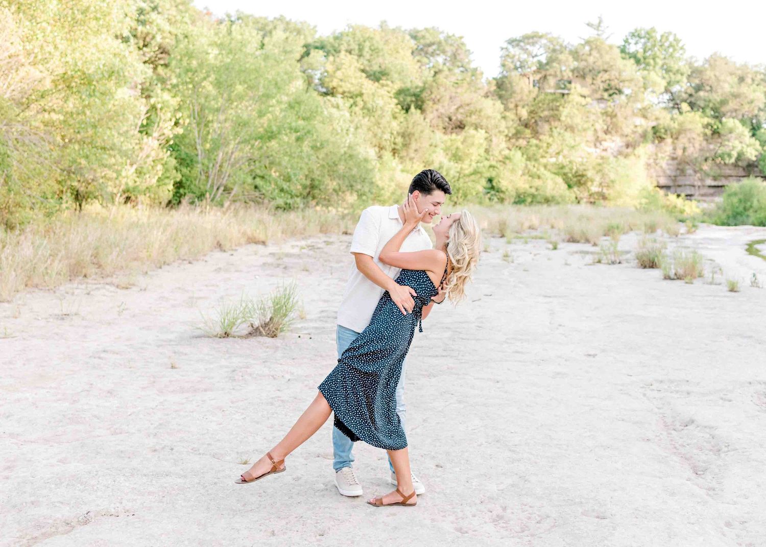 Couple dancing together on a sandy riverbank surrounded by green trees.
