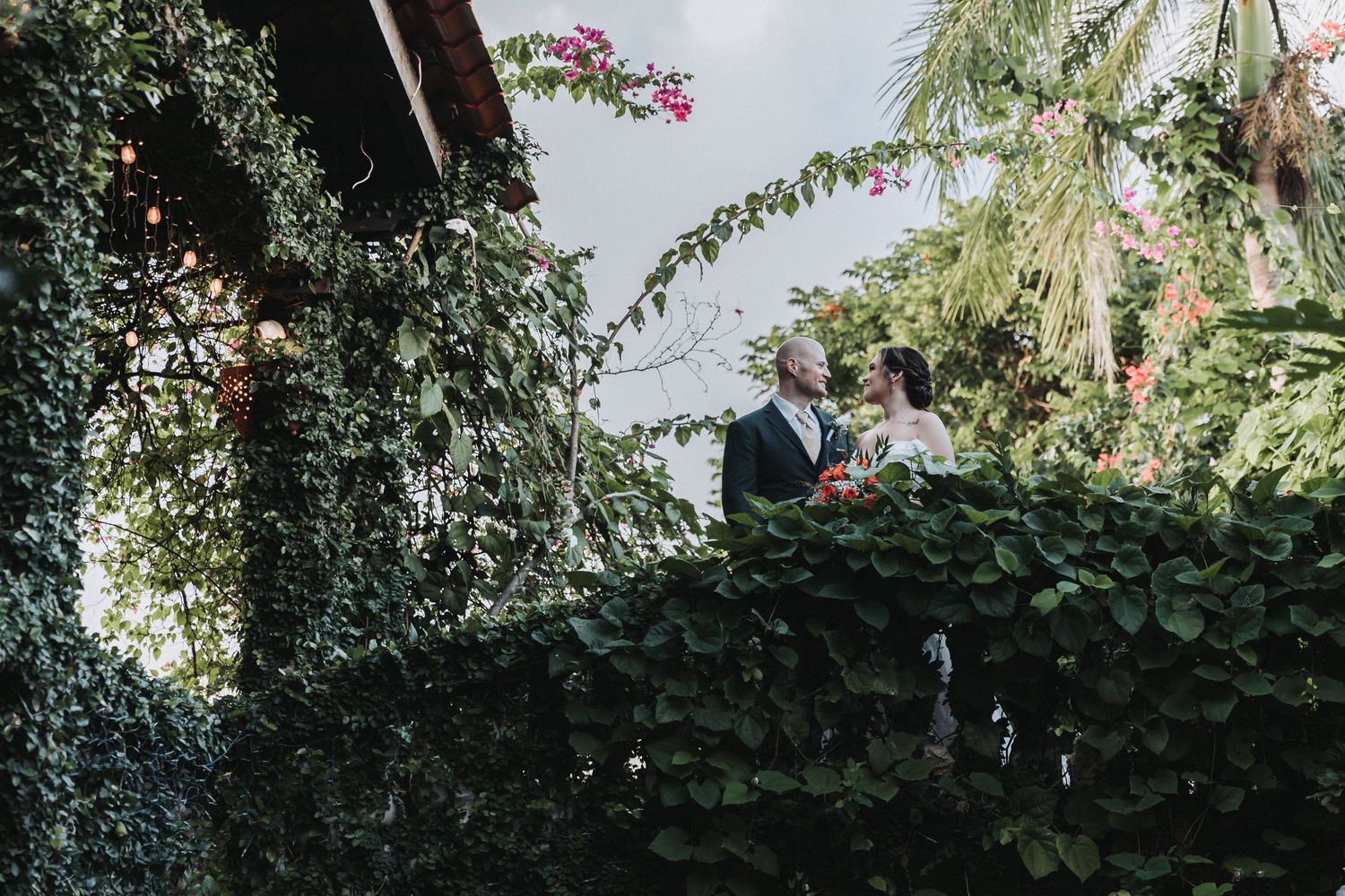 A couple stands together on an ivy-covered balcony surrounded by lush greenery and flowers.