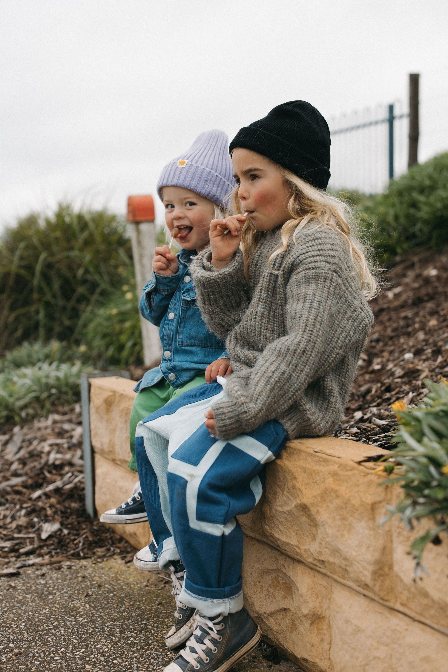 Two people in winter attire sitting together on a stone wall wearing knit sweaters and beanies on an overcast day.