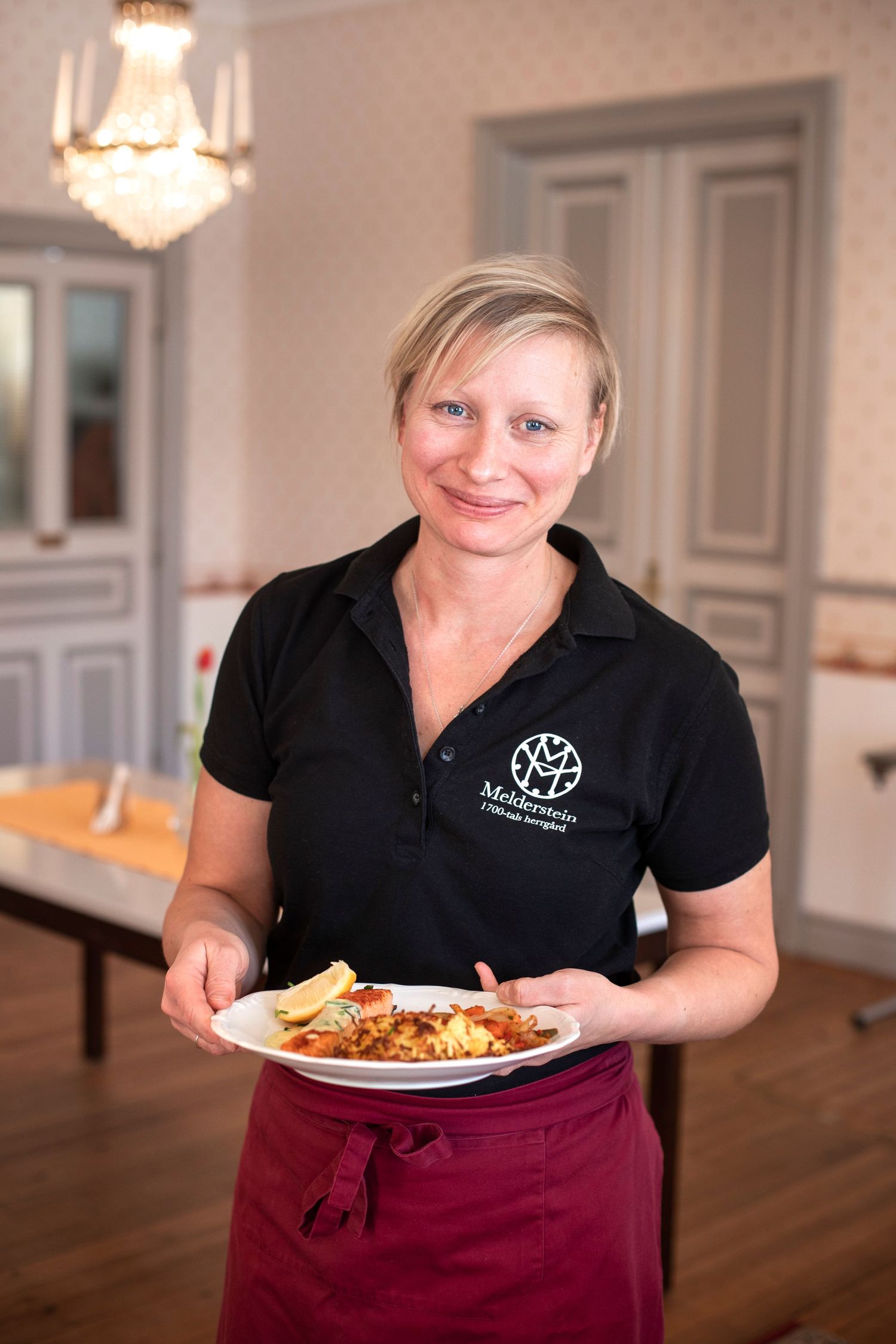 Smiling waitress in black uniform holding a plate of food in an elegant dining room.