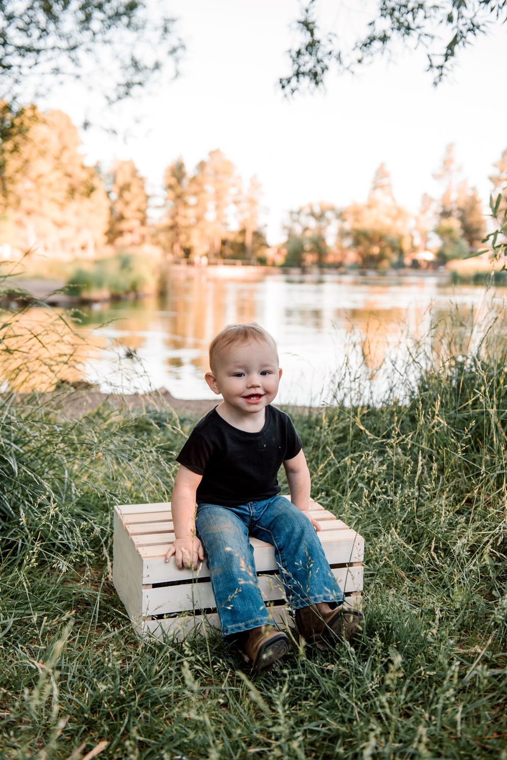 Small child sits on wooden bench surrounded by tall grass near a lake during golden hour.