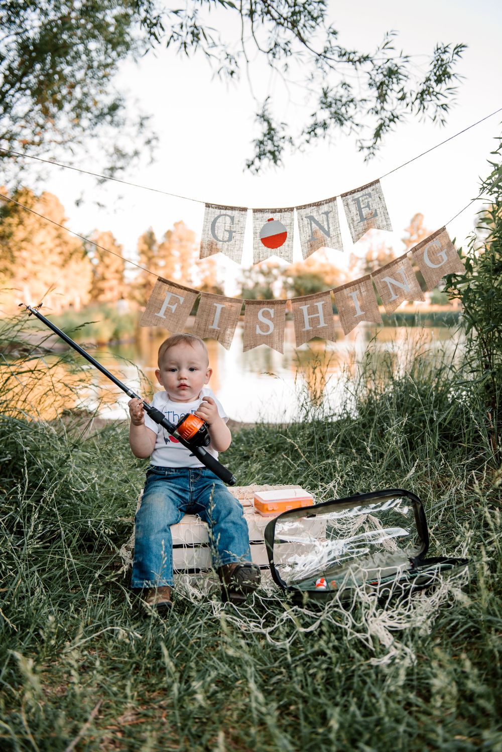 Child playing with fishing gear on grassy lakeside with decorative banner overhead.