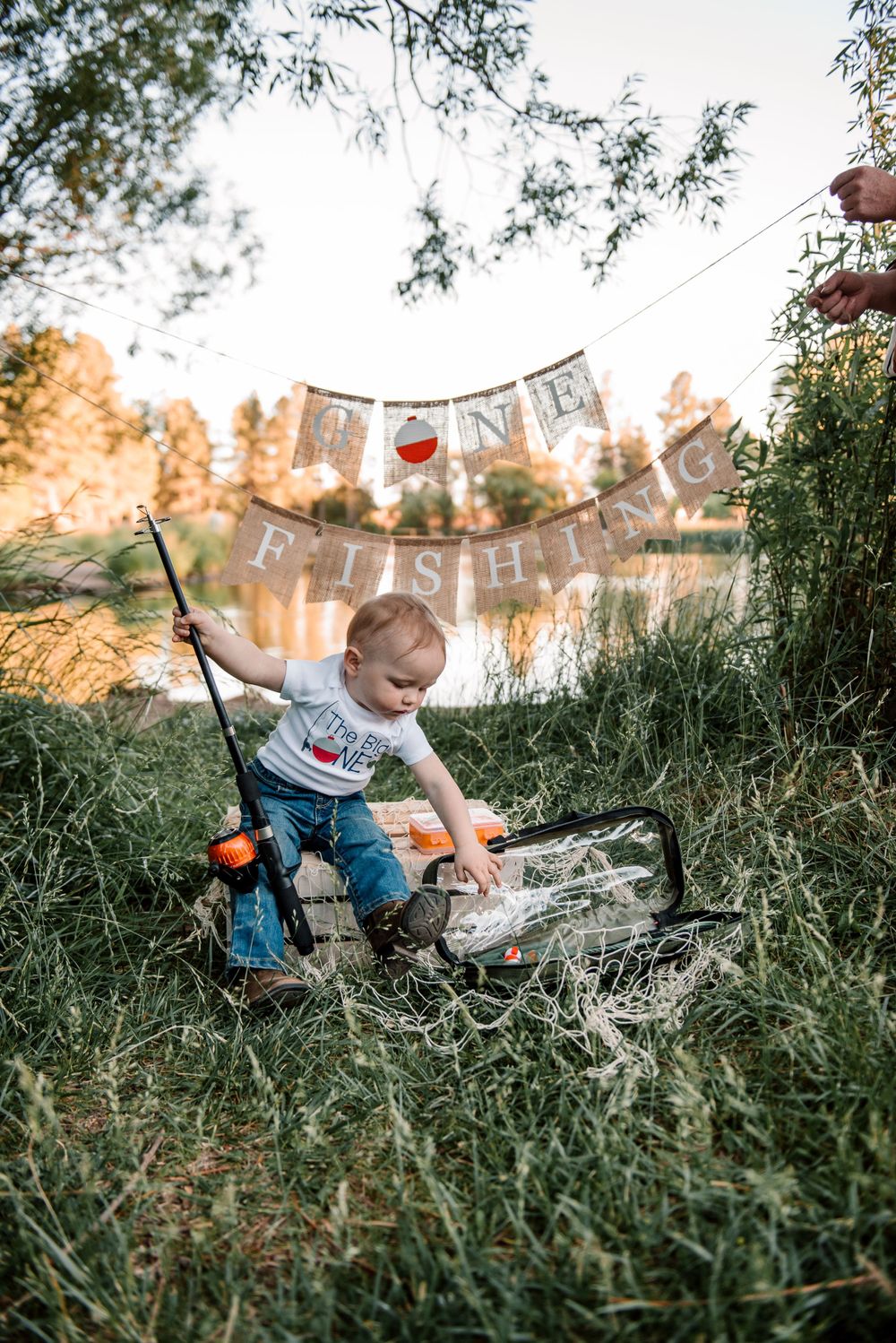 Toddler explores fishing equipment while sitting in tall grass with festive bunting above.