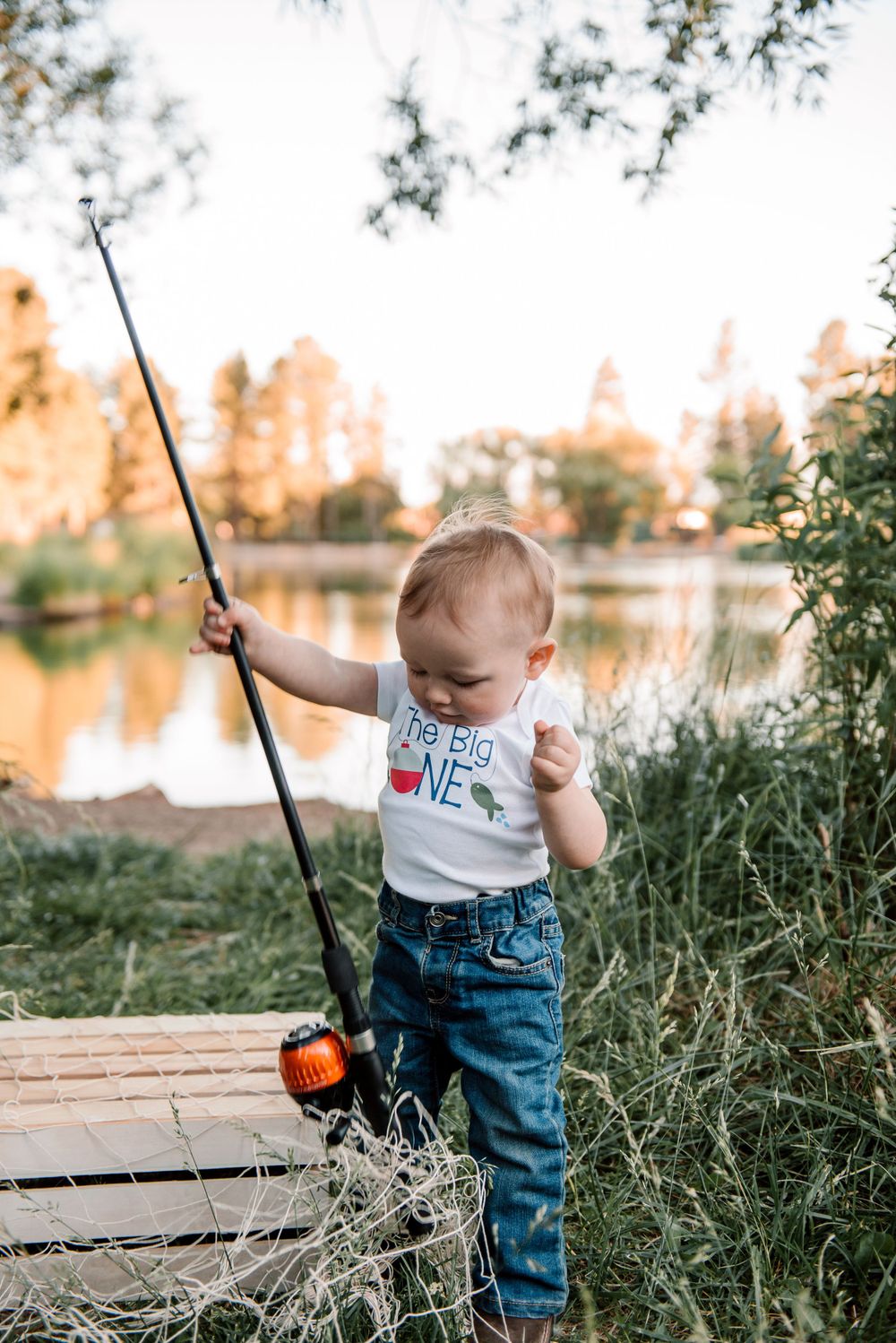Child holds fishing rod while playing outdoors by lake during sunset.