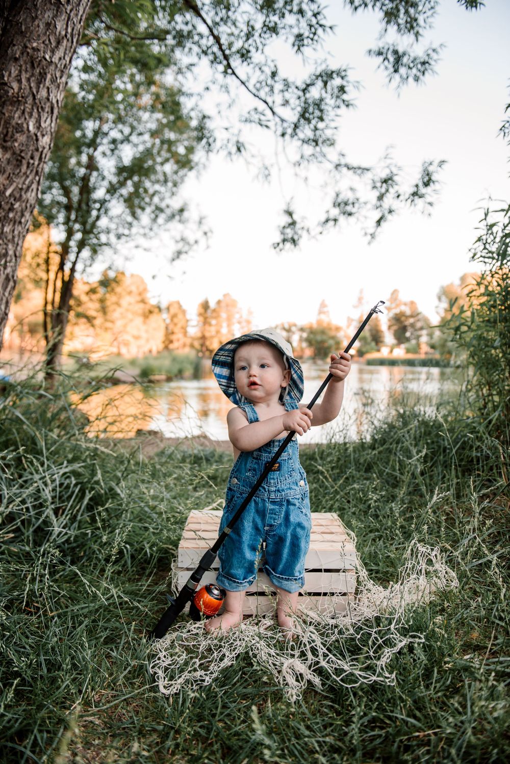 Toddler plays with fishing pole while sitting in tall grass near water.