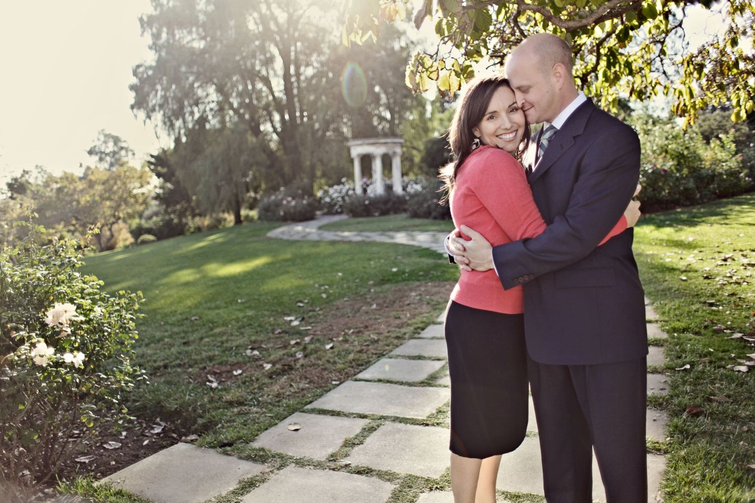 Romantic couple embracing on a garden path with a gazebo visible in the background.