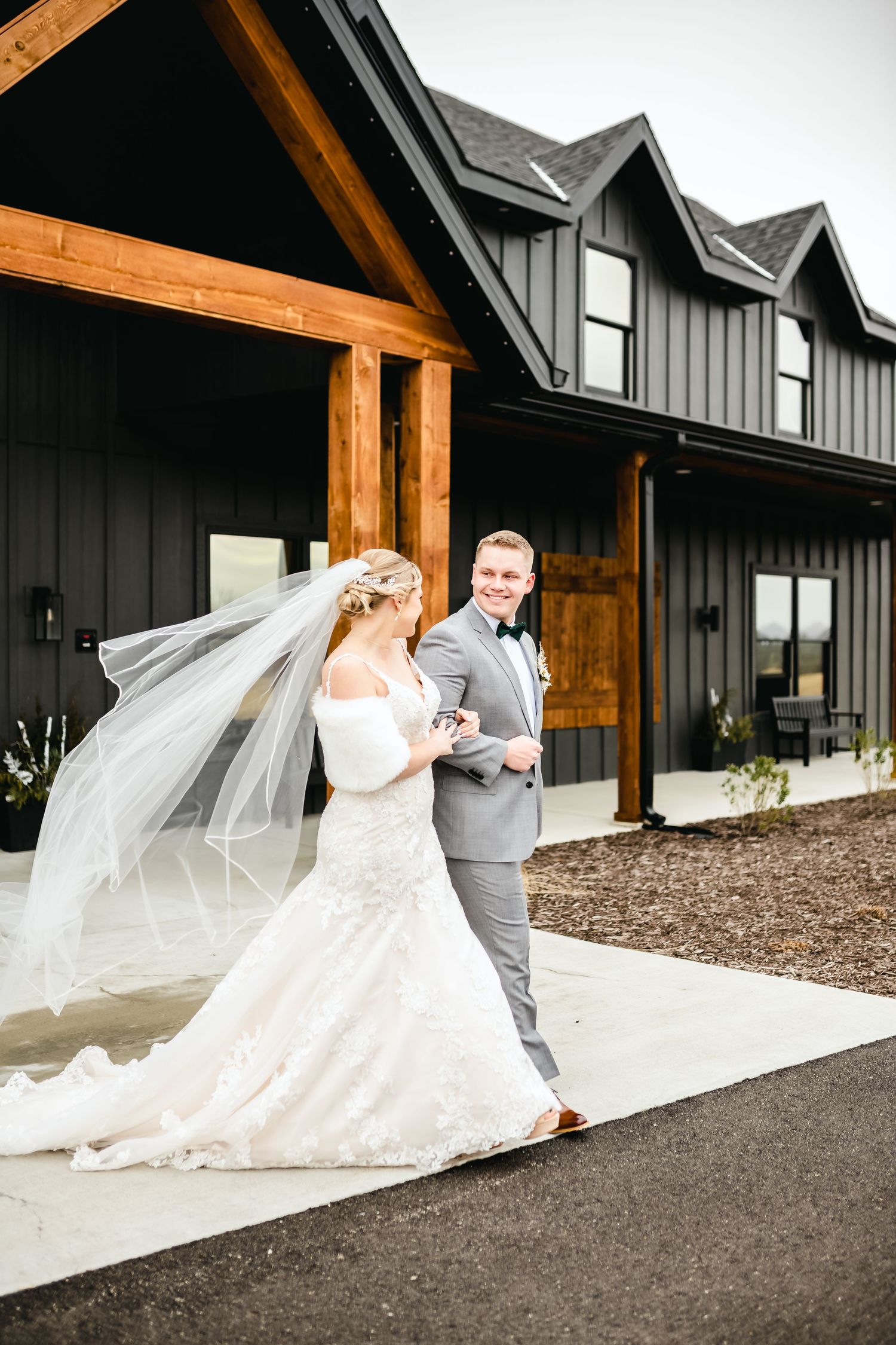 A flowing white wedding dress and veil captured in motion against a modern black and wood building.