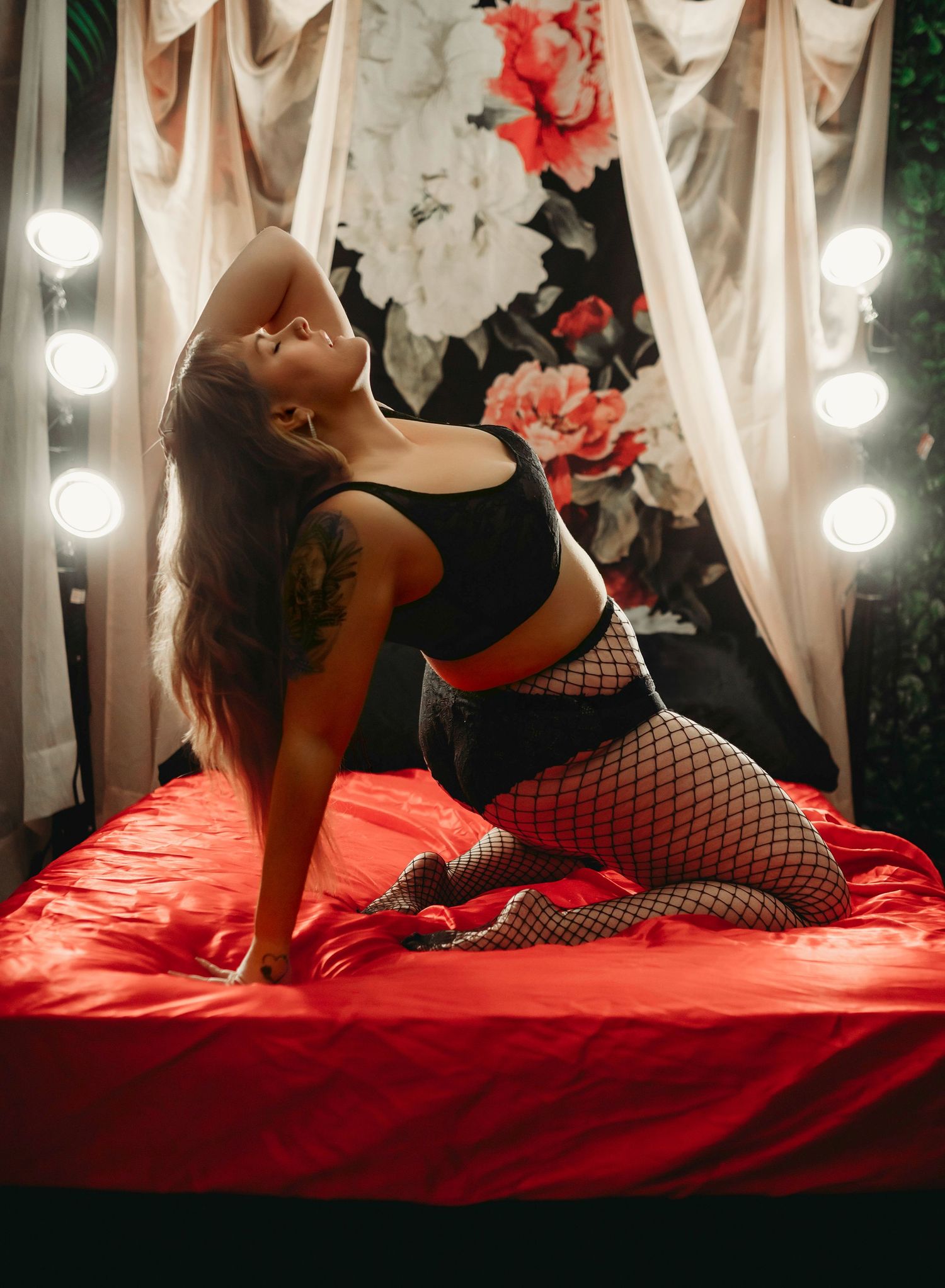 Artistic boudoir photo with red floral backdrop, dramatic lighting and fishnet stockings on red bedding.