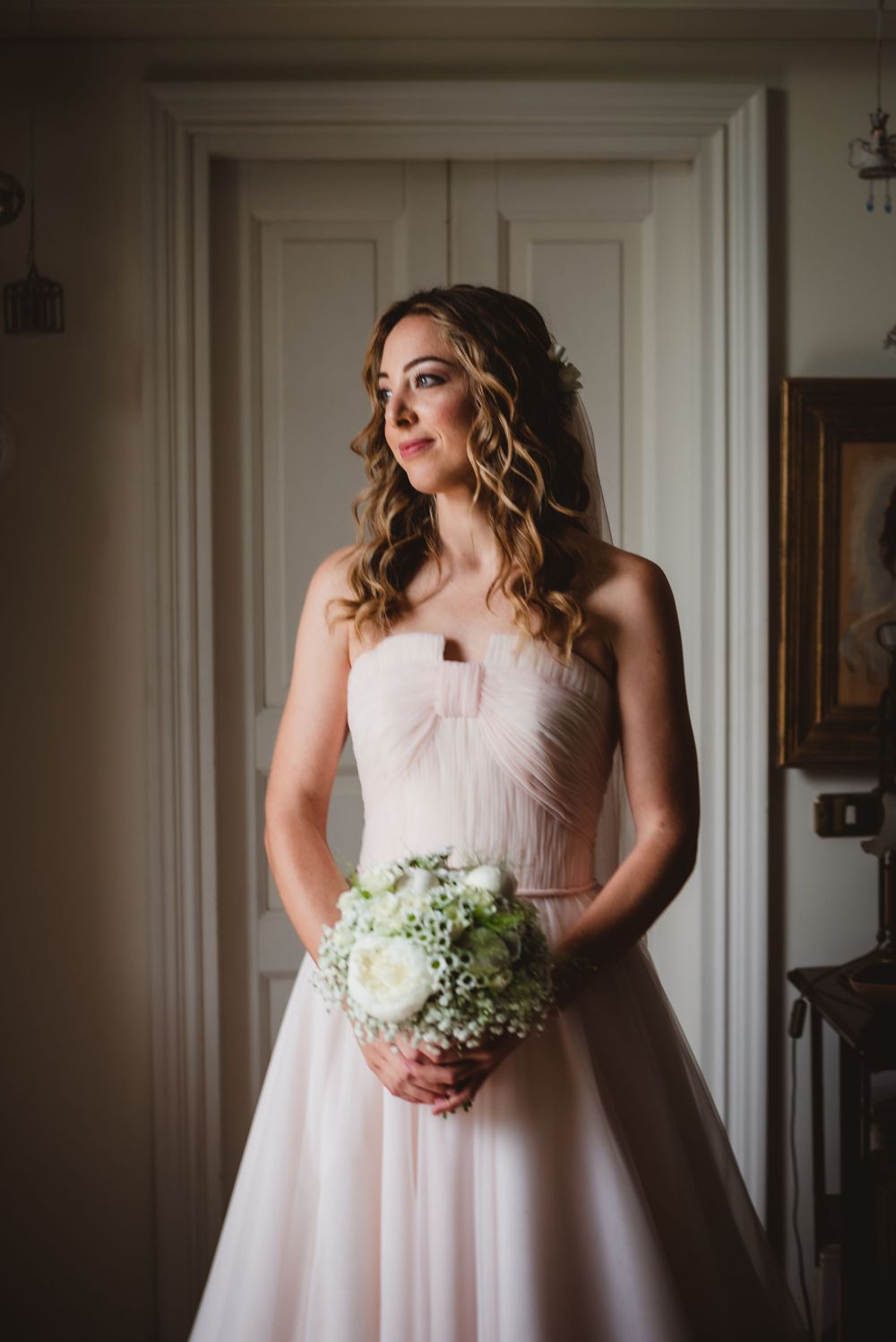 Bride in a blush pink gown holds white bouquet while posing near white doors.