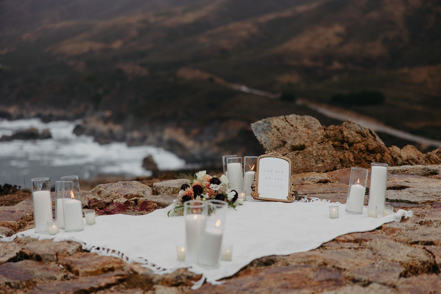 Romantic setup with candles and white fabric on rocky coastal cliffs at dusk.