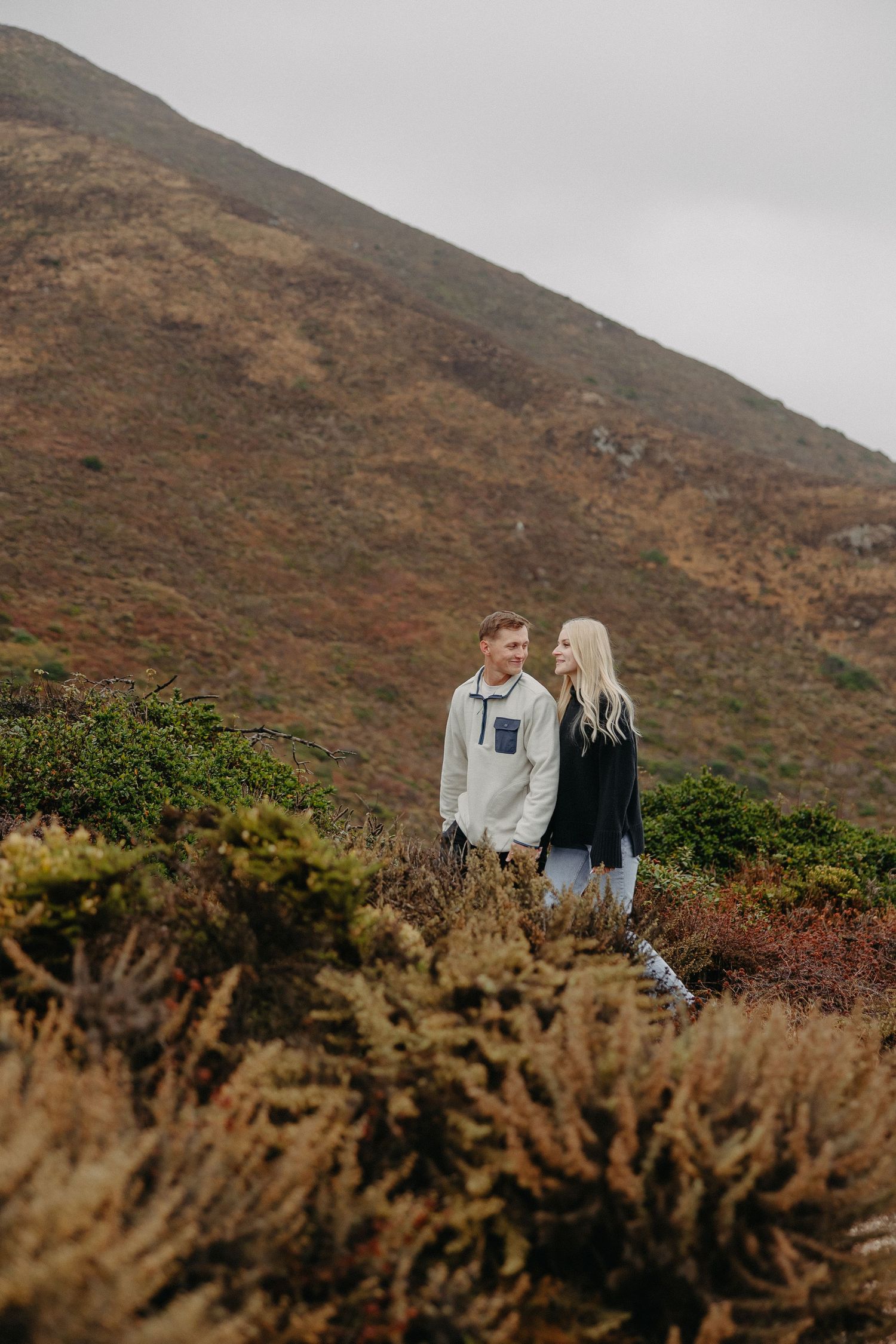 A couple stands together on a rugged hillside overlooking a coastal landscape on an overcast day.