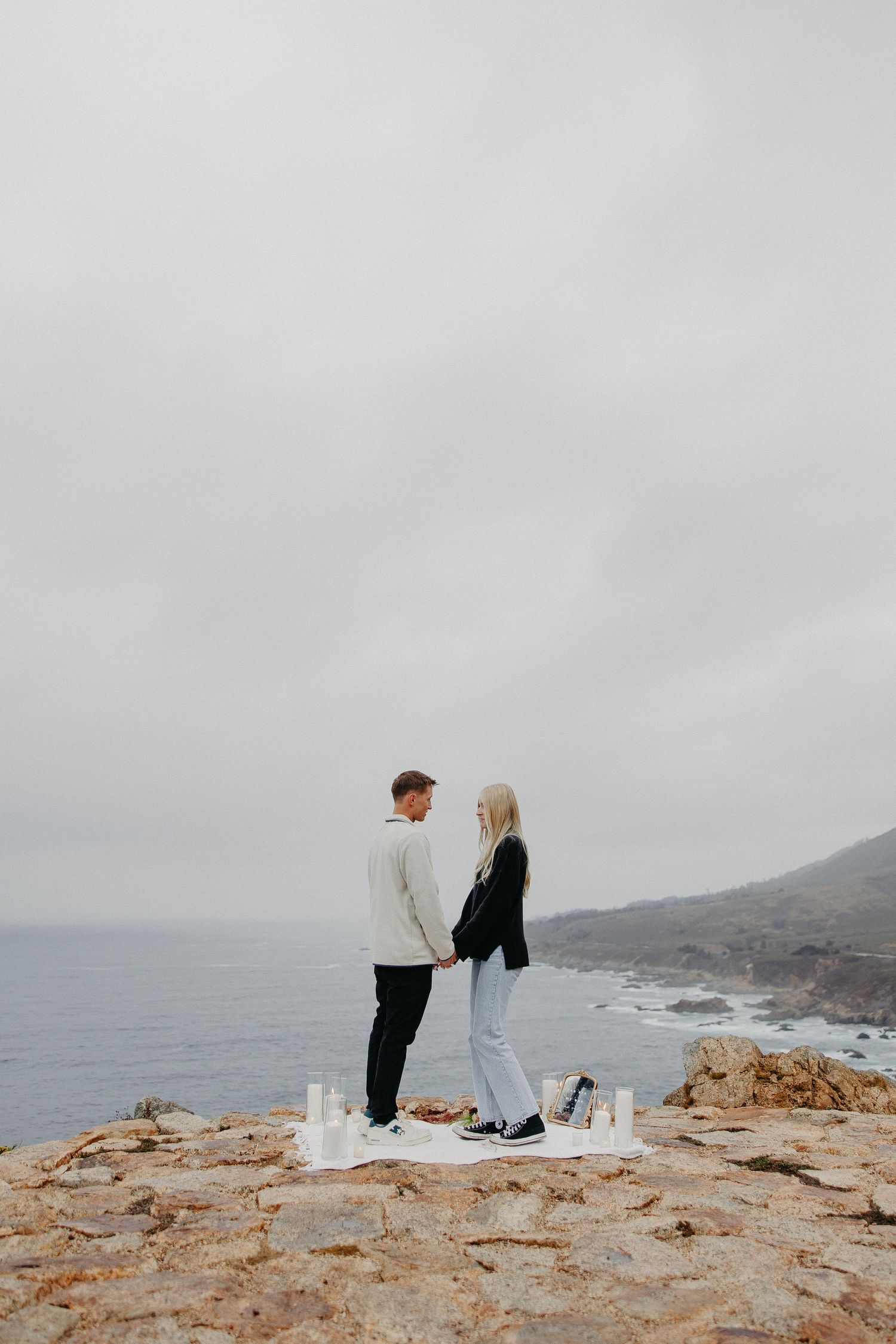 Two people stand on a rocky cliff overlooking the ocean with a moody grey sky backdrop.