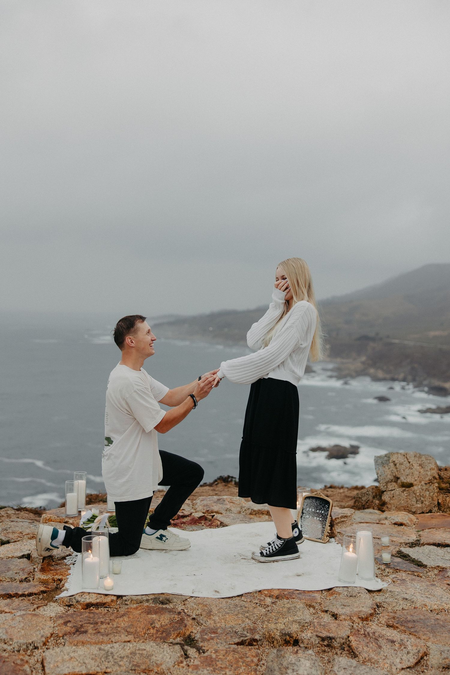 Romantic proposal scene with couple on blanket surrounded by candles on coastal cliff.