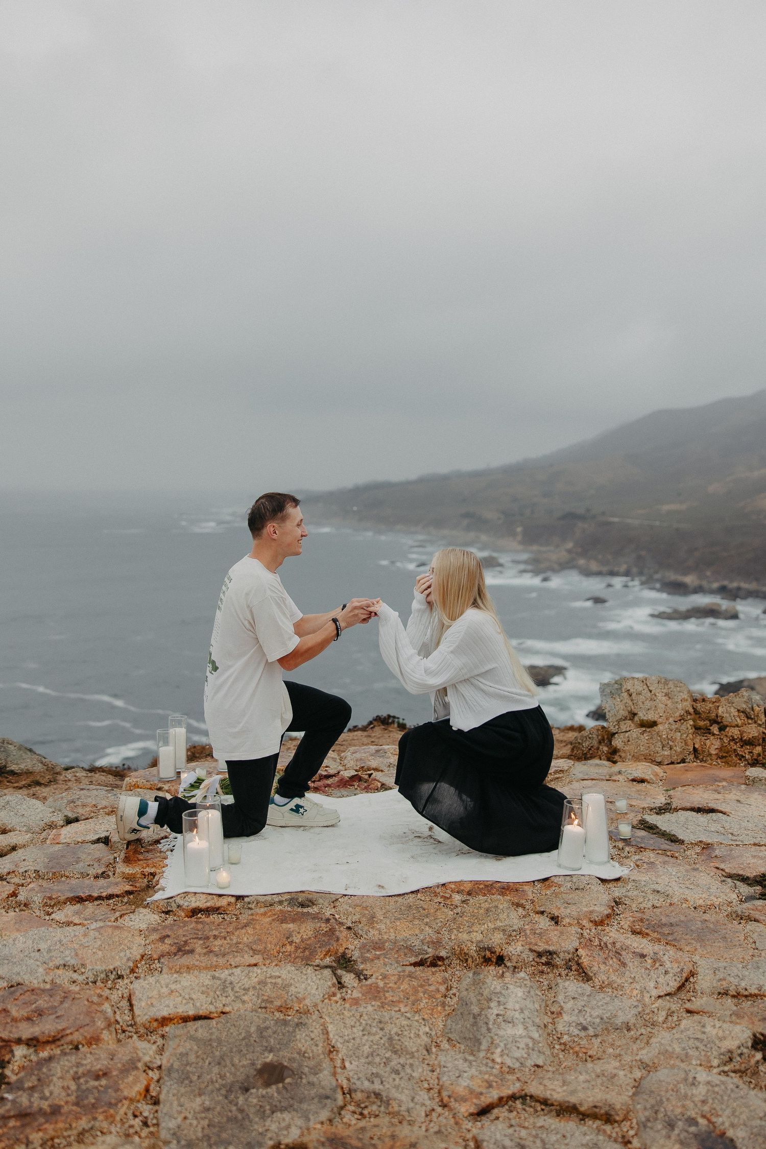 A romantic proposal moment on a rocky coastal cliff overlooking the ocean during an overcast day.