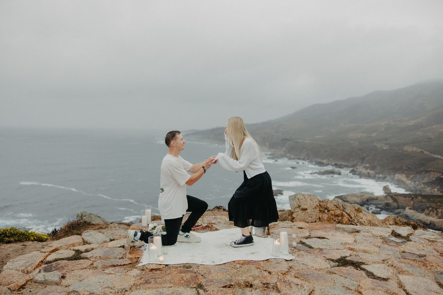A proposal setup on coastal rocks with white blanket and couple in casual attire.