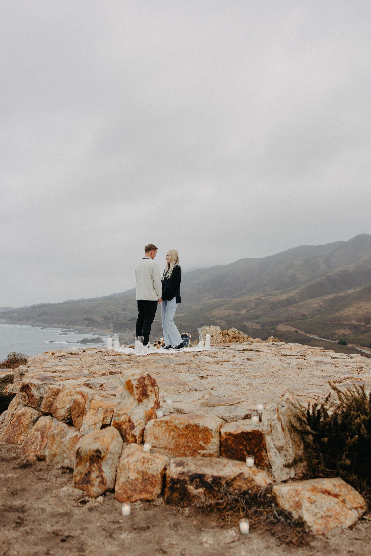 A couple shares a moment on a rocky coastal overlook with dramatic mountain views.