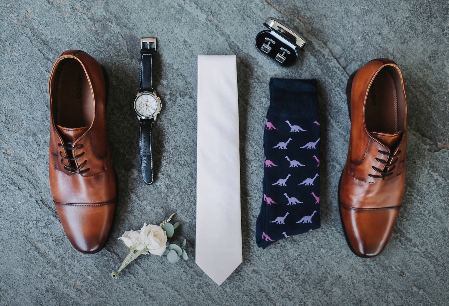 Brown leather oxford shoes arranged symmetrically with tie, watch and patterned socks on gray background.