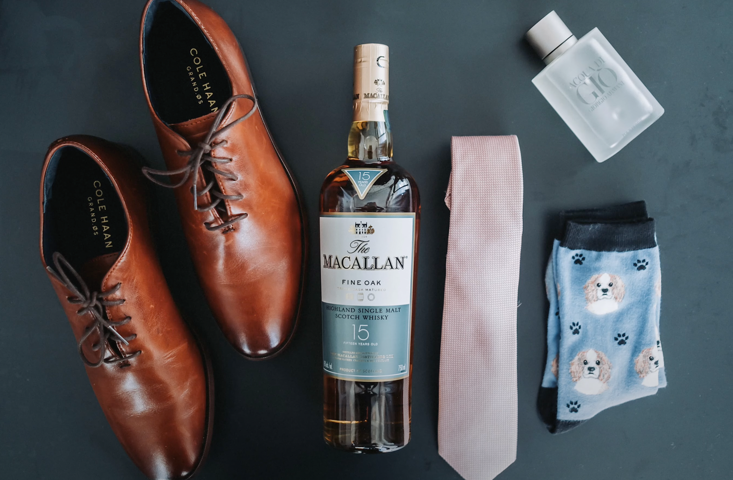 Brown leather dress shoes arranged with Macallan whisky, pink tie, and patterned socks on wooden surface.