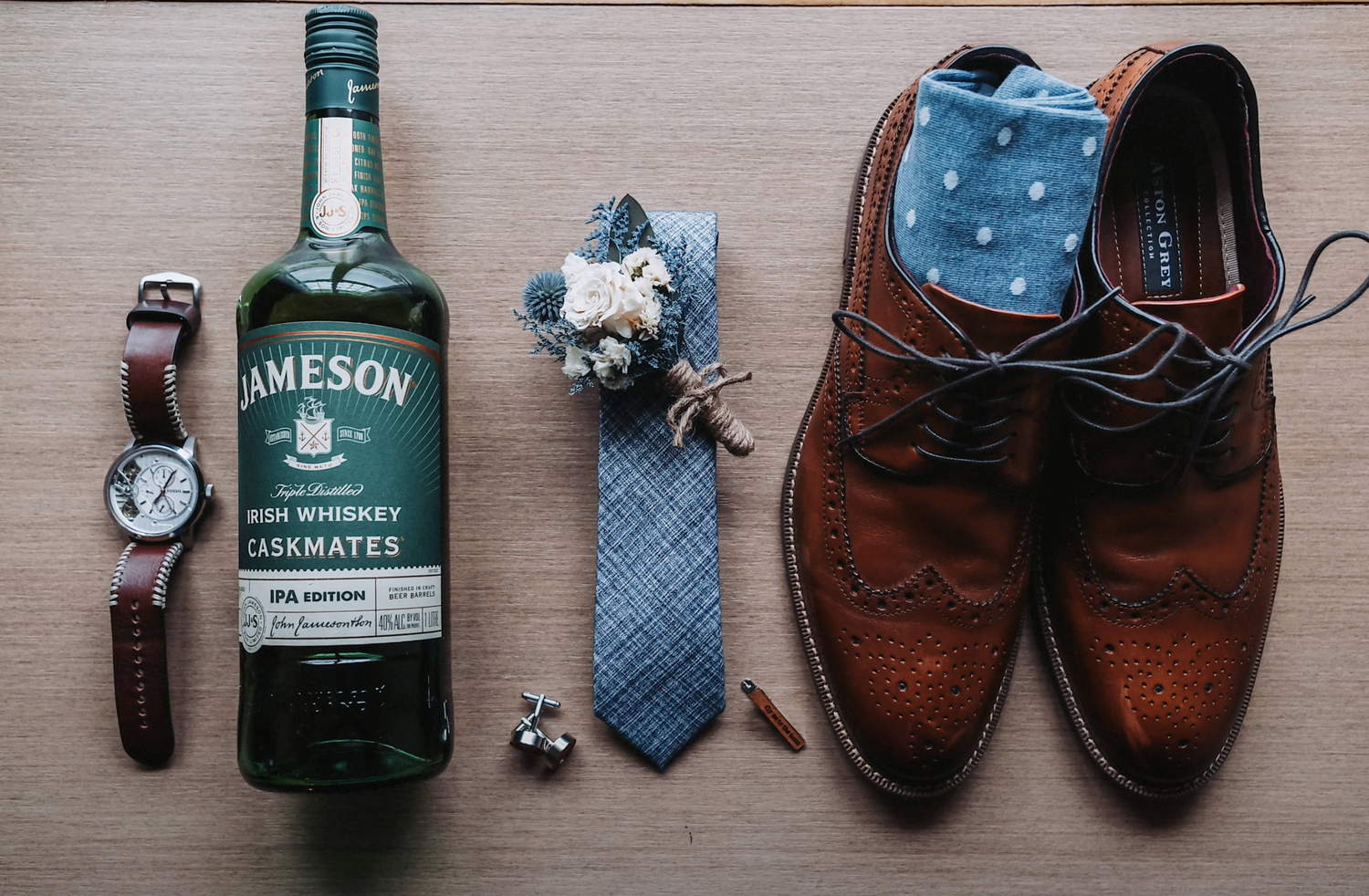Jameson whiskey bottle displayed with brown oxford shoes, wristwatch, and blue tie on wooden surface.