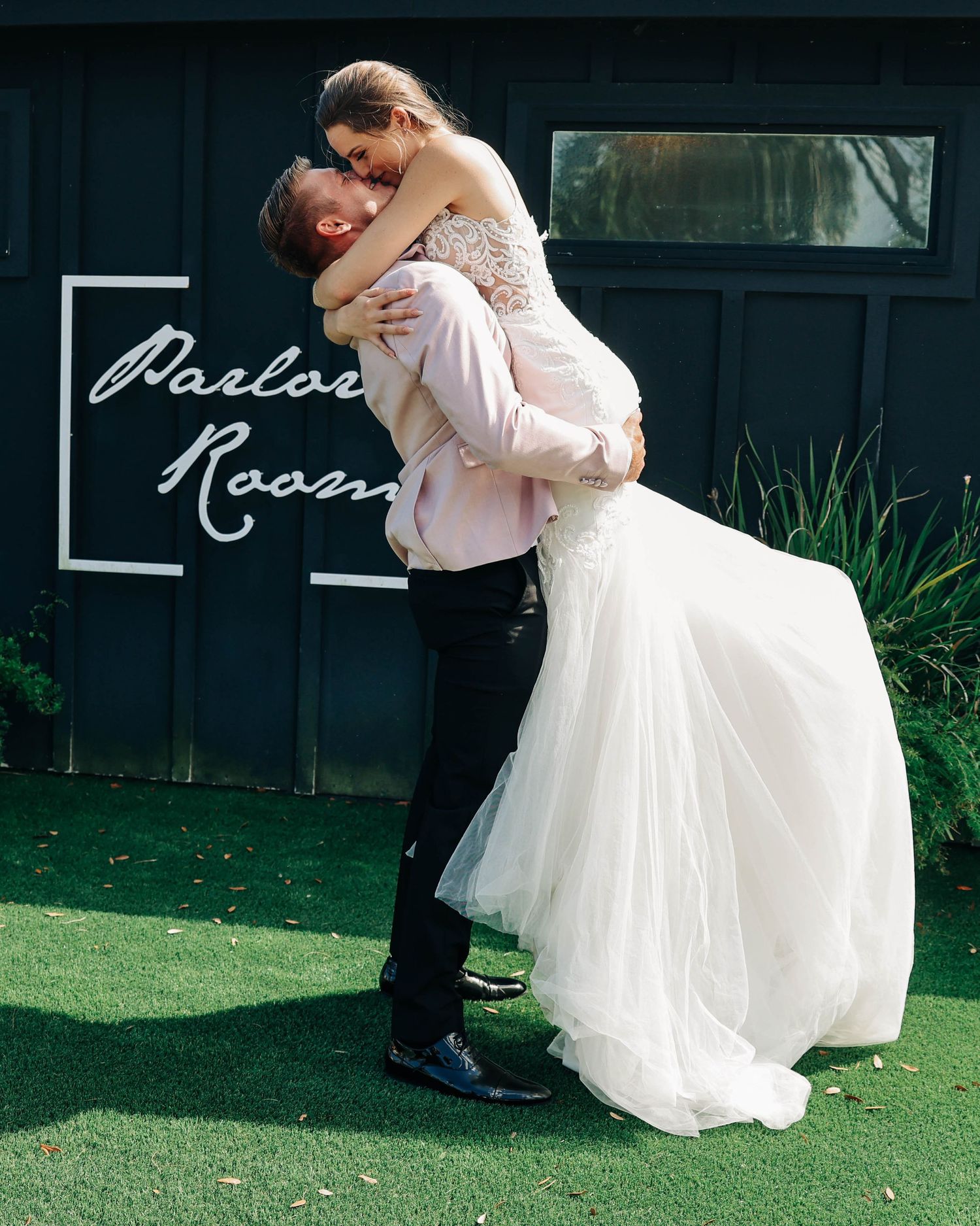 Romantic wedding couple embrace outside venue with modern signage and greenery.