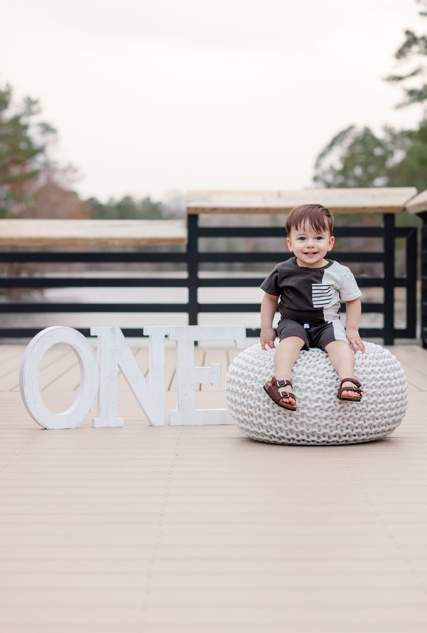 Child sitting on white knitted pouf next to 'ONE' letters on outdoor deck.