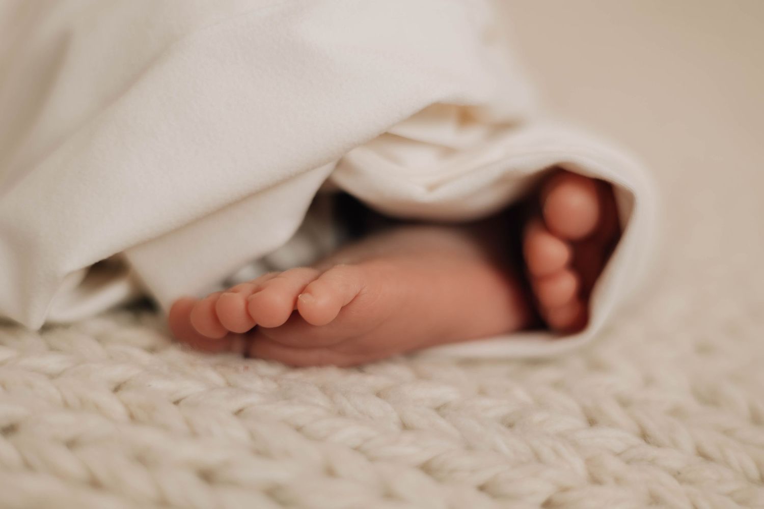 Close-up of newborn baby feet peeking out from under a white blanket on a knit surface.