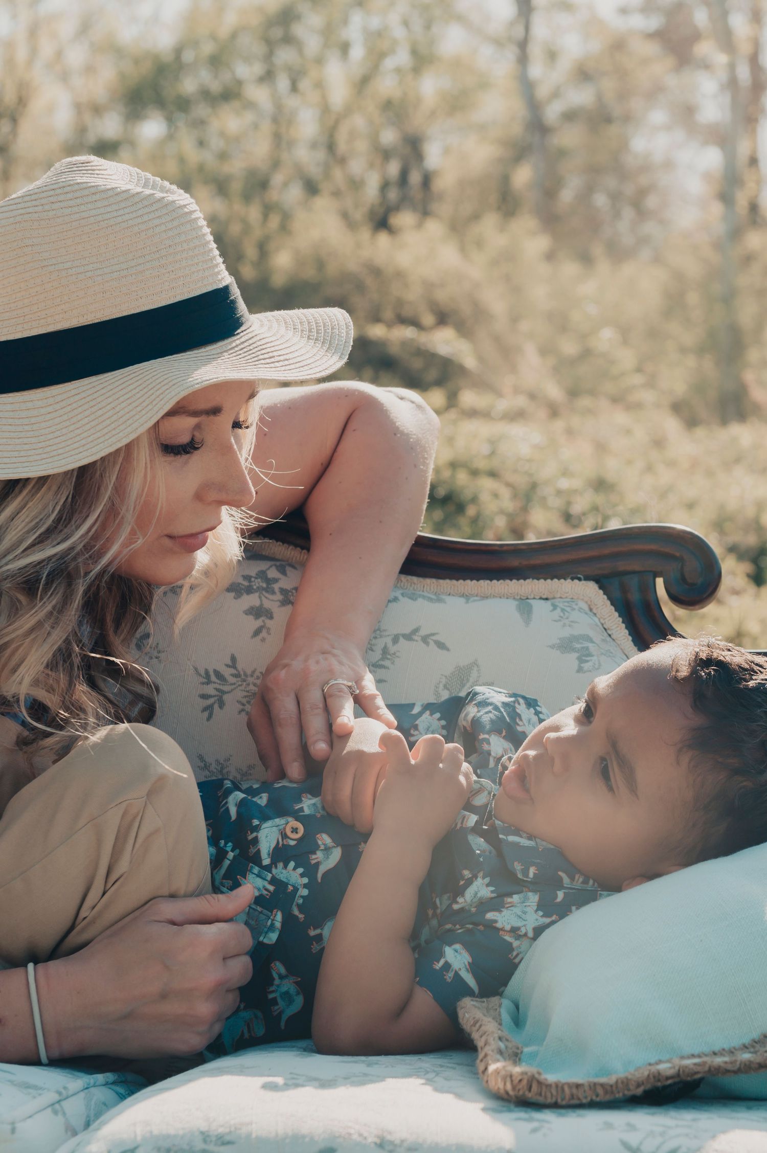Tender outdoor photo shoot showing a lady in a sun hat lying on a blue floral blanket in natural sunlight.