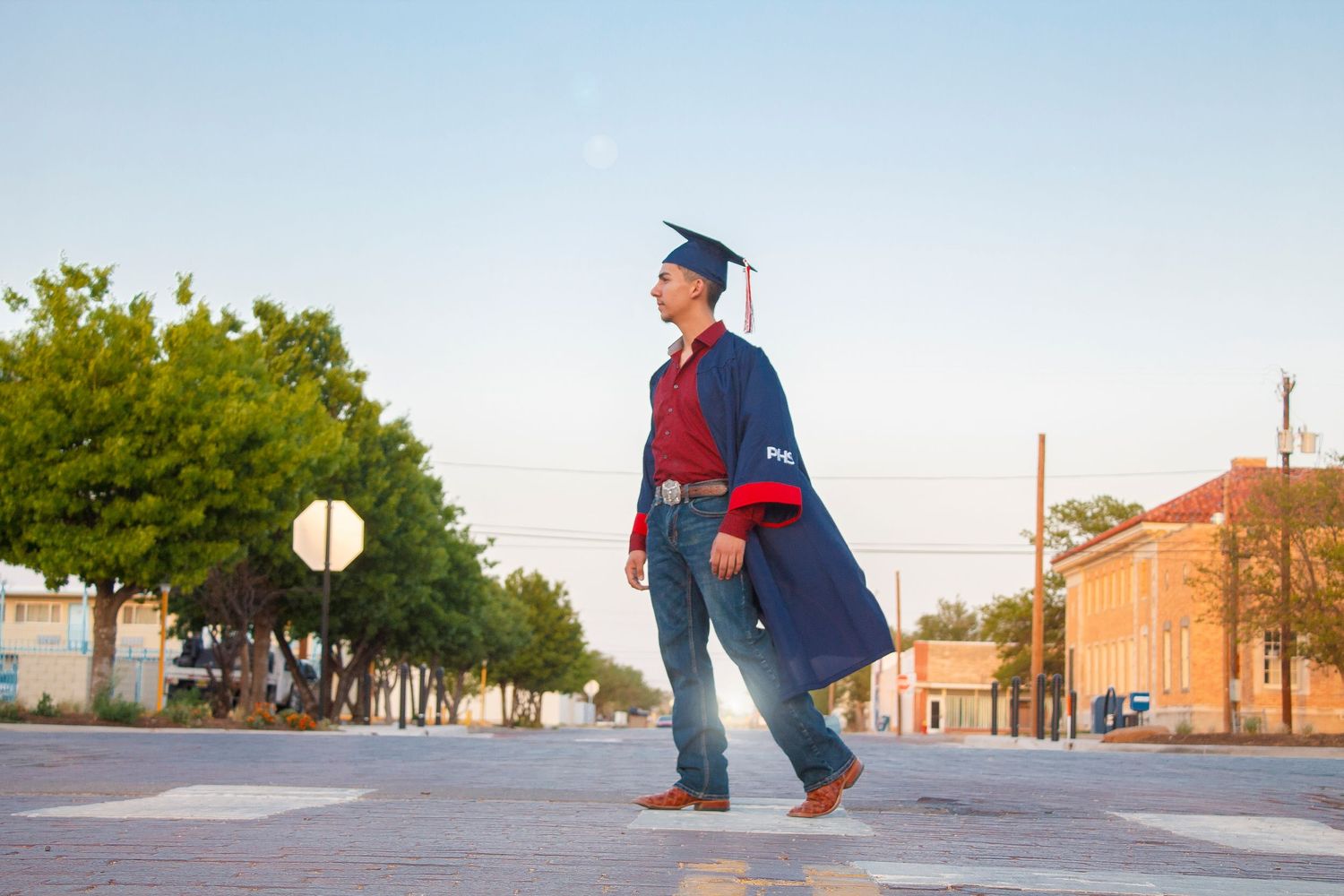 A graduate in cap and gown poses on an empty street at sunset.