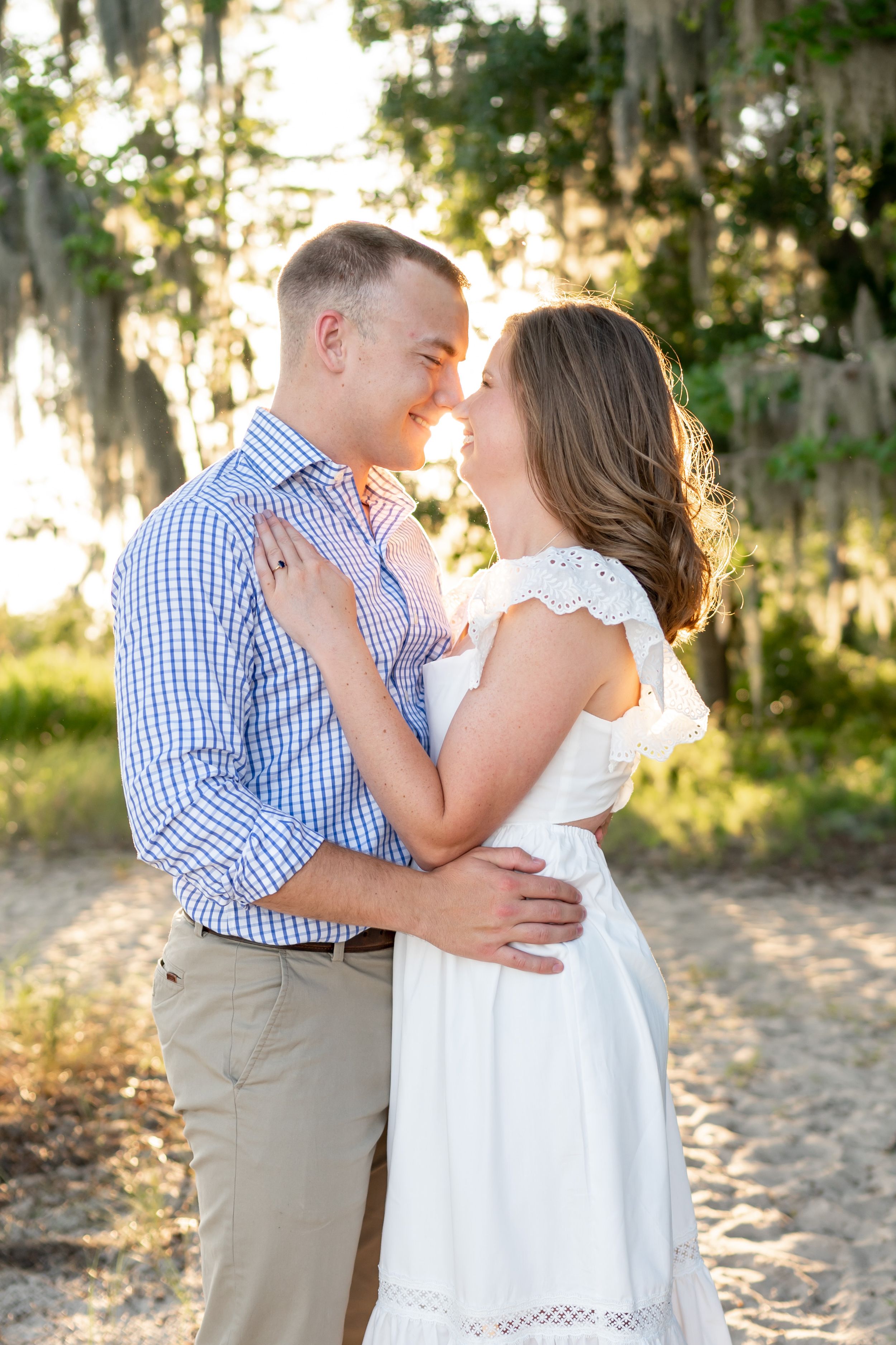 Couple embraces in romantic pose during sunset with Spanish moss hanging in background.