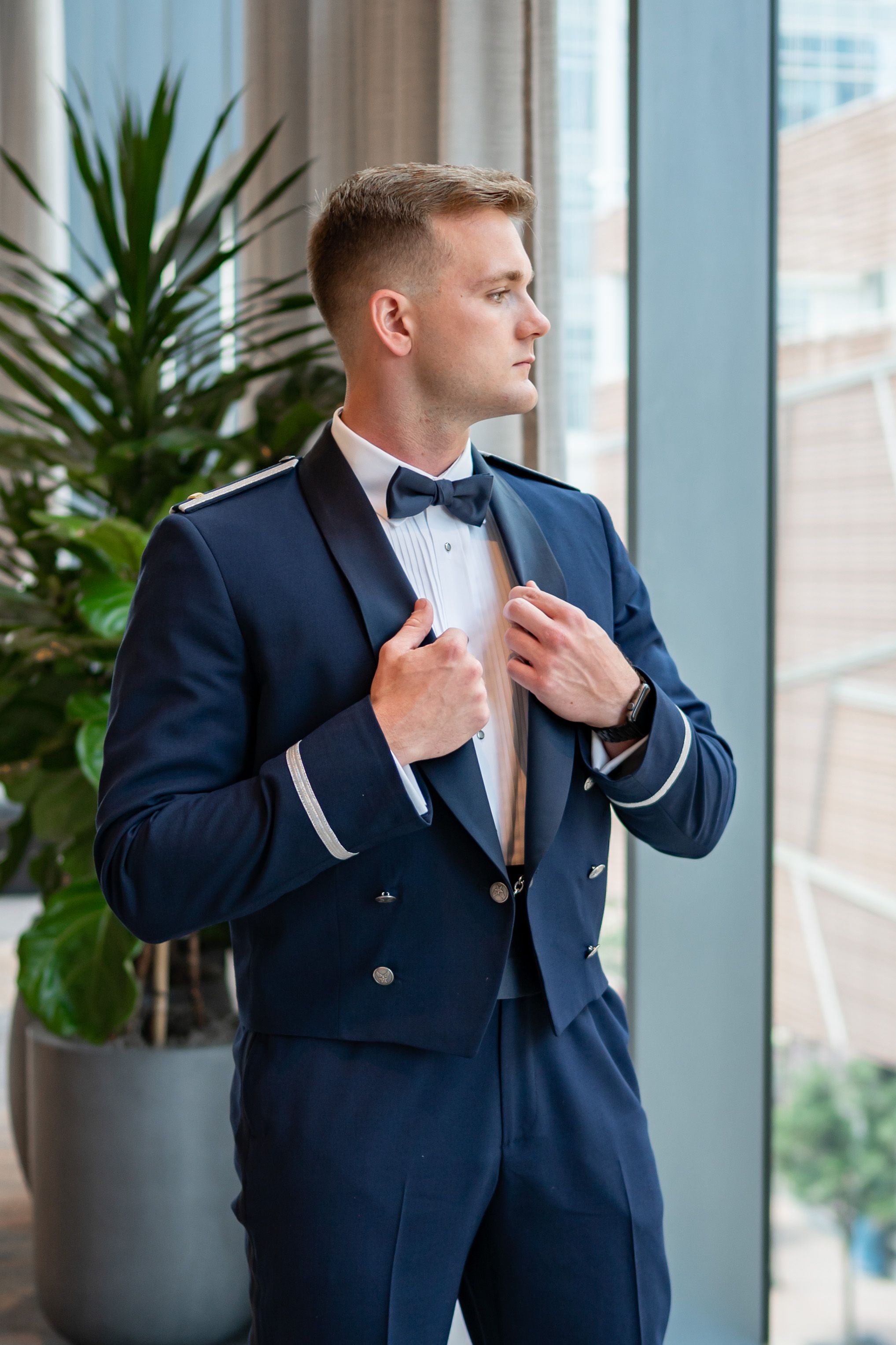 A formal portrait of someone in a navy blue suit adjusting their bowtie near a window.