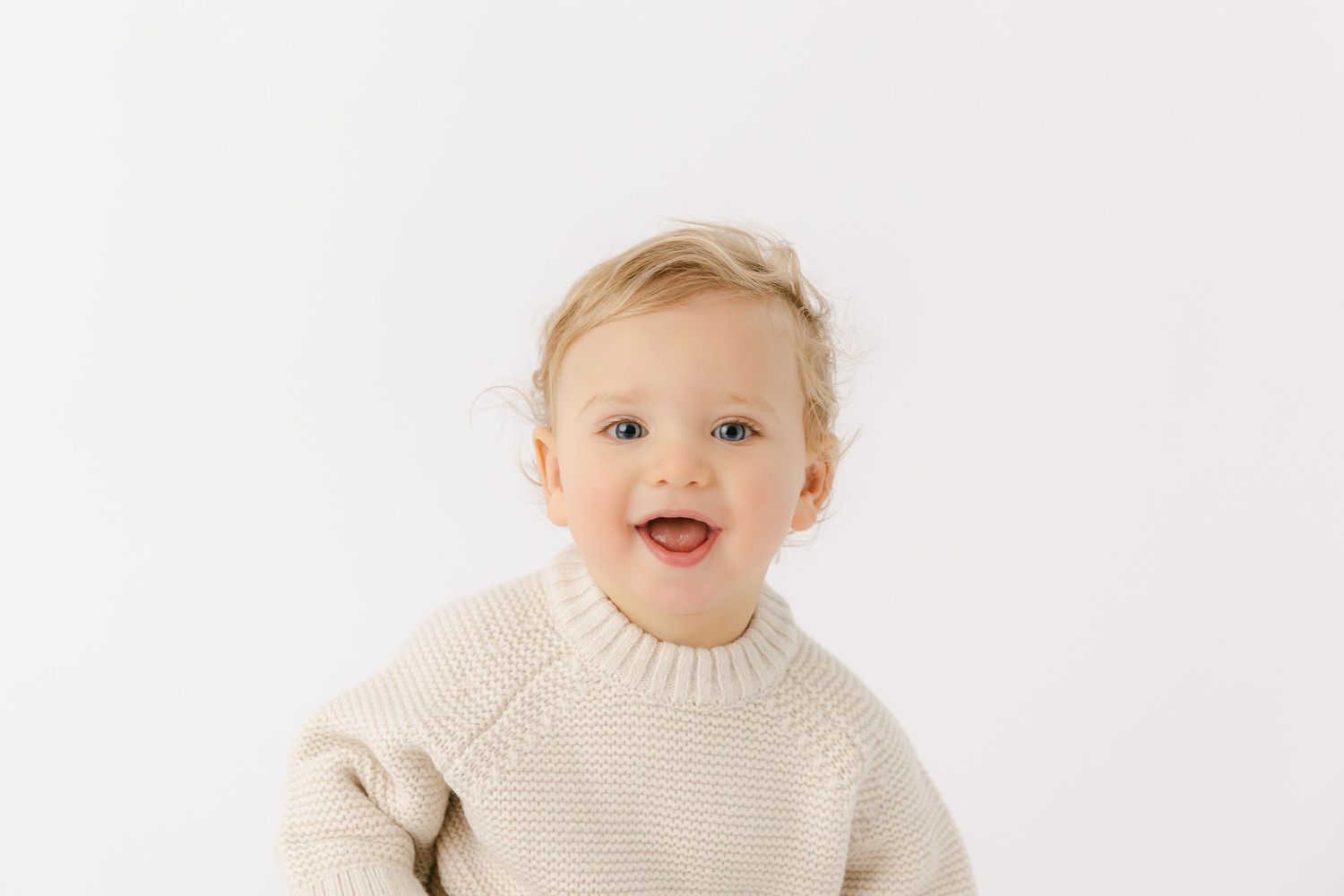 Close up portrait of baby with blonde hair smiling. Baby is wearing a cream knit sweater.