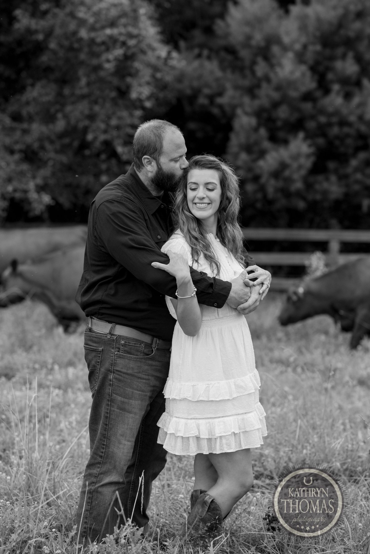 A couple shares a tender moment while standing in a pasture with horses in the background.