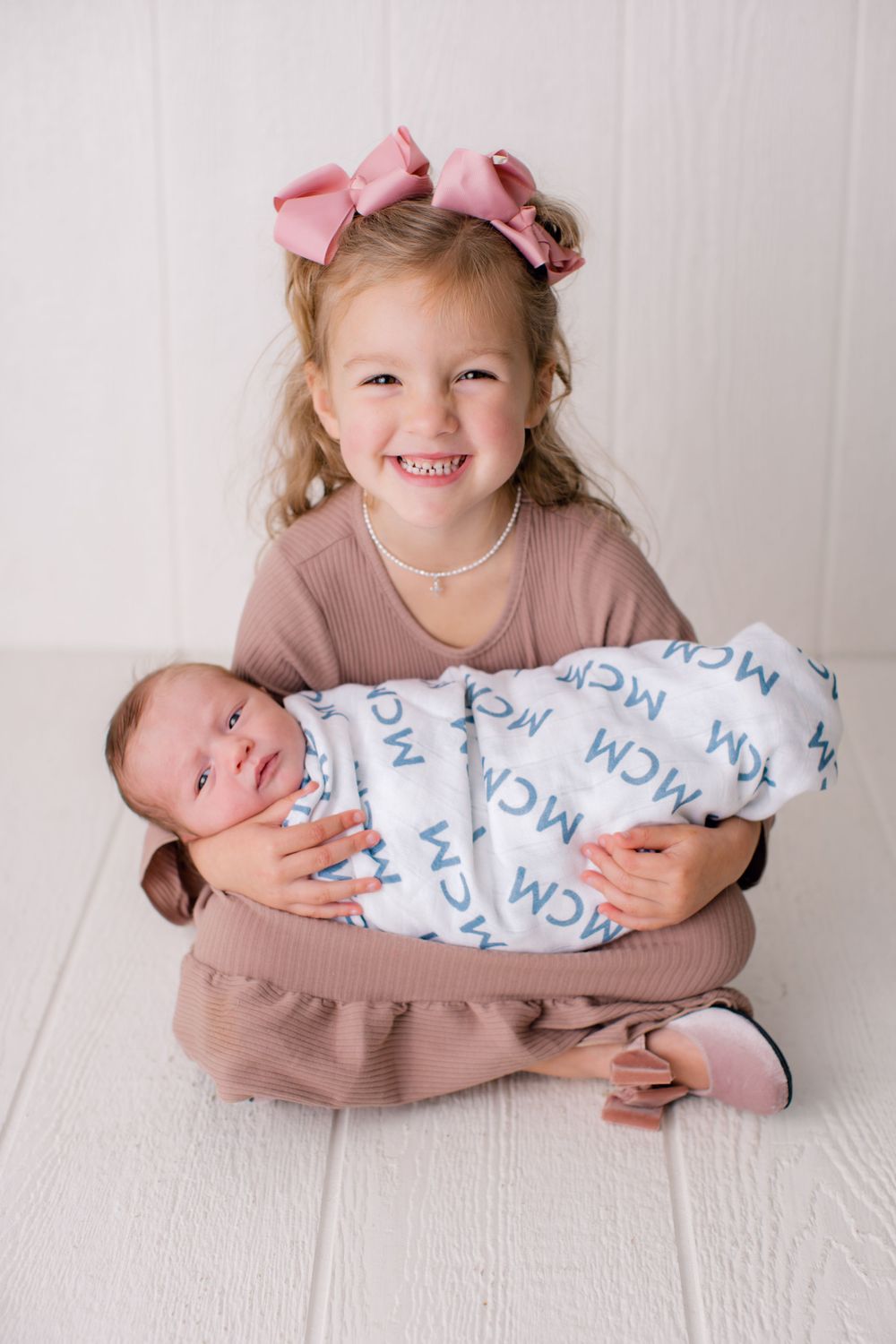 A young child wearing pink bows holds a swaddled newborn baby during a professional photoshoot.