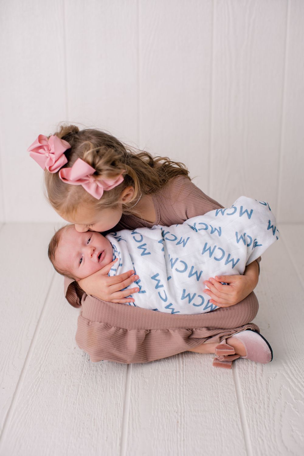 A sweet sibling moment captured as a young child cuddles a swaddled newborn on white flooring.