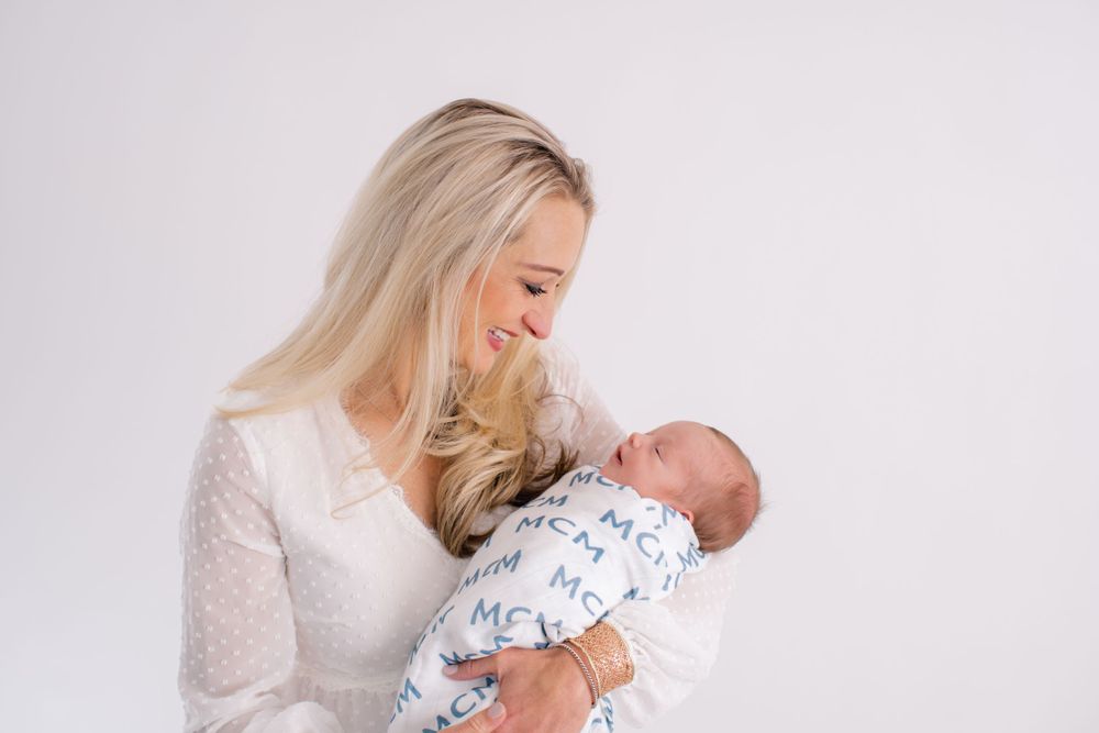 Mother in white dress lovingly holds swaddled newborn baby against light background.