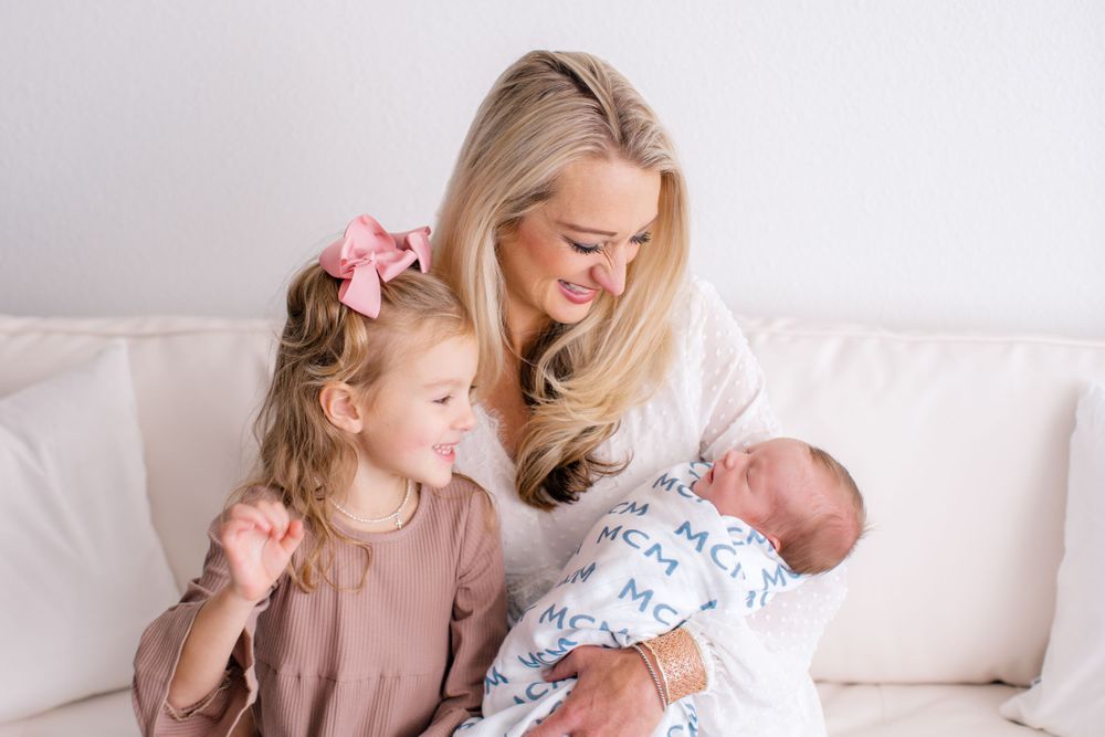 Sweet family scene of mother and daughter looking at newborn baby on white couch.