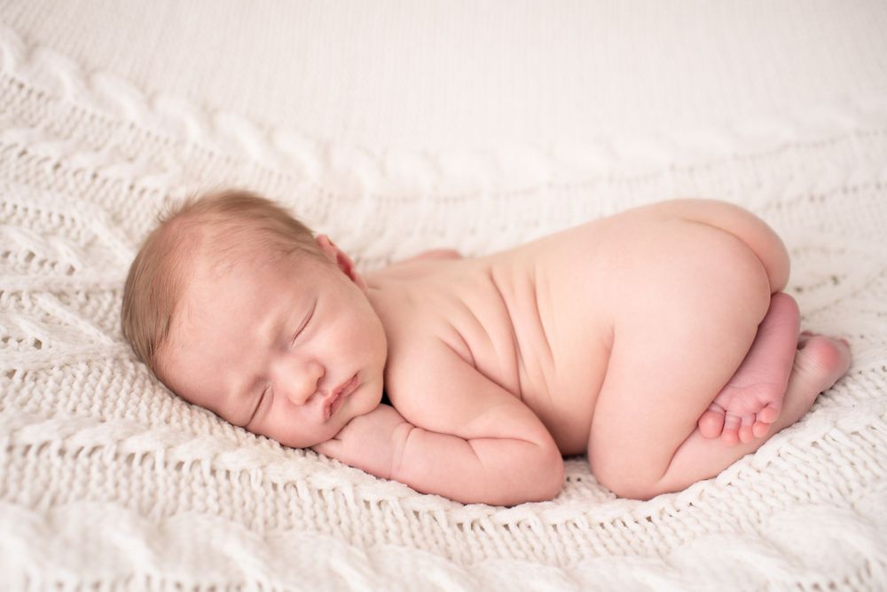 Sleeping newborn baby curled up on white knitted blanket during studio photography session.